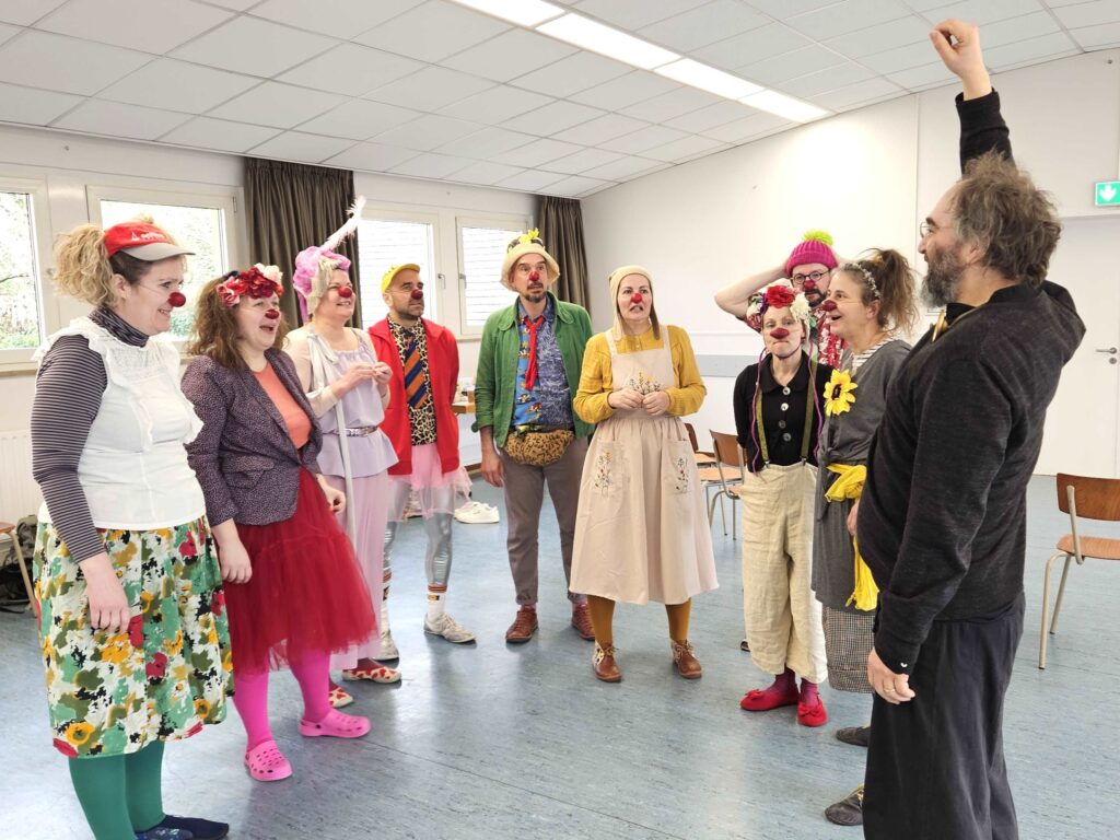 Un groupe de personnes déguisées en clowns se tient en cercle dans une pièce lumineuse, écoutant un instructeur au bras levé. Elles portent des costumes colorés, des nez rouges et des visages expressifs, et semblent engagées et joyeuses.