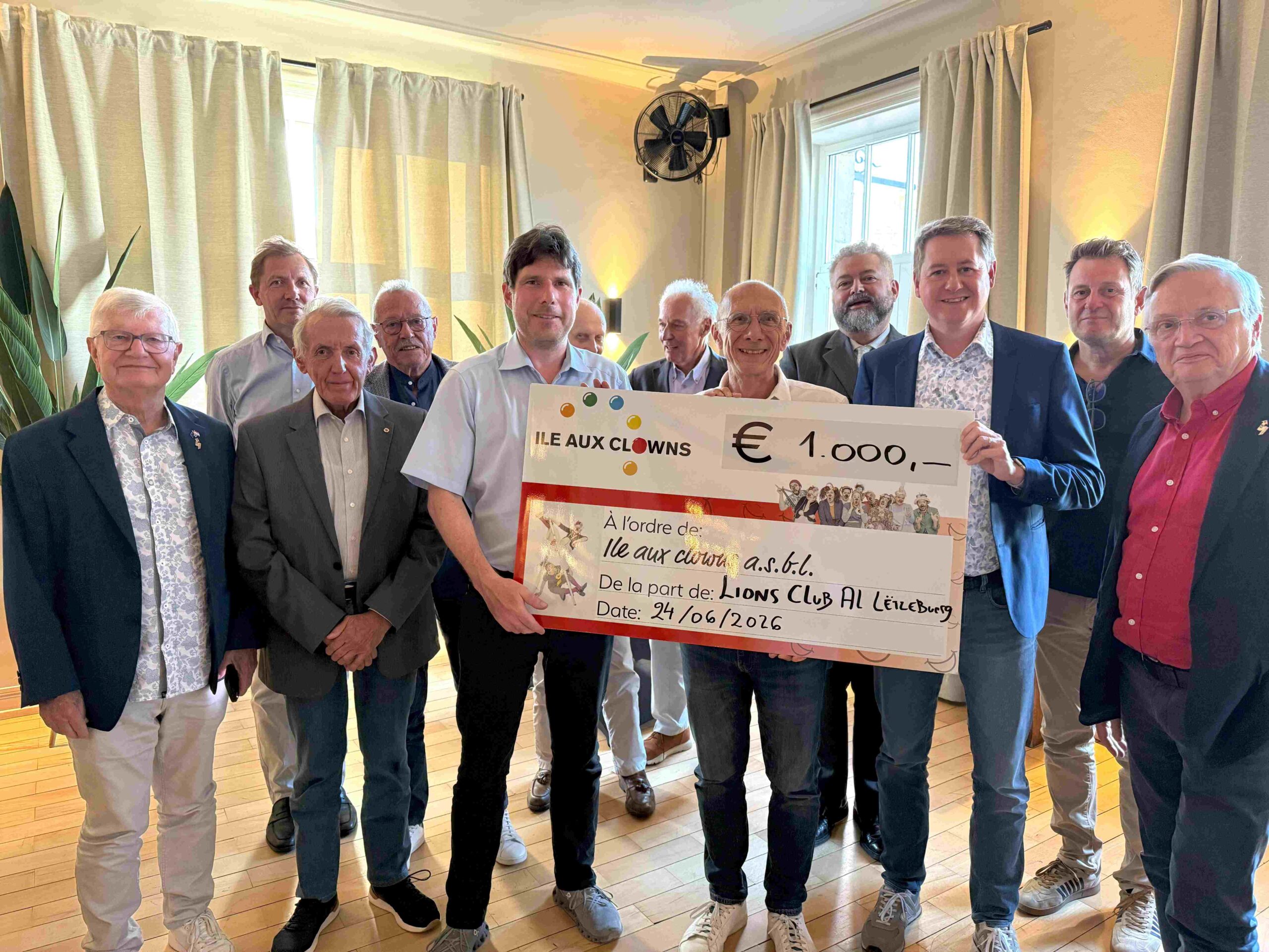 Un groupe de onze hommes se tient à l'intérieur, souriant et tenant un gros chèque de cérémonie de 1 000 euros à l'ordre de l'Île aux Clowns, émis par le Lions Club AL Esch/Alzette. La pièce est bien éclairée par de grandes fenêtres et des rideaux clairs.