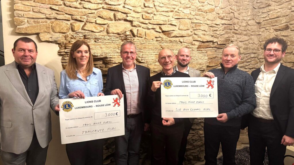 Sept personnes se tiennent à l'intérieur, devant un mur de pierre, souriantes et tenant deux grands chèques de 3 000 euros chacun du Lions Club Luxembourg-Roude Léiw, libellés à l'ordre de deux organisations différentes.