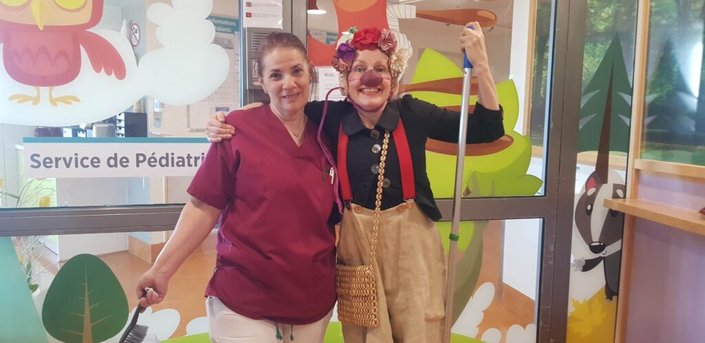 Deux femmes sourient à l'intérieur : l'une en blouse médicale tenant une tapette à mouches se tient à côté d'une femme habillée en clown avec un nez rouge, des lunettes, une couronne de fleurs et un balai. Elles se trouvent devant l'entrée d'un service de pédiatrie coloré.