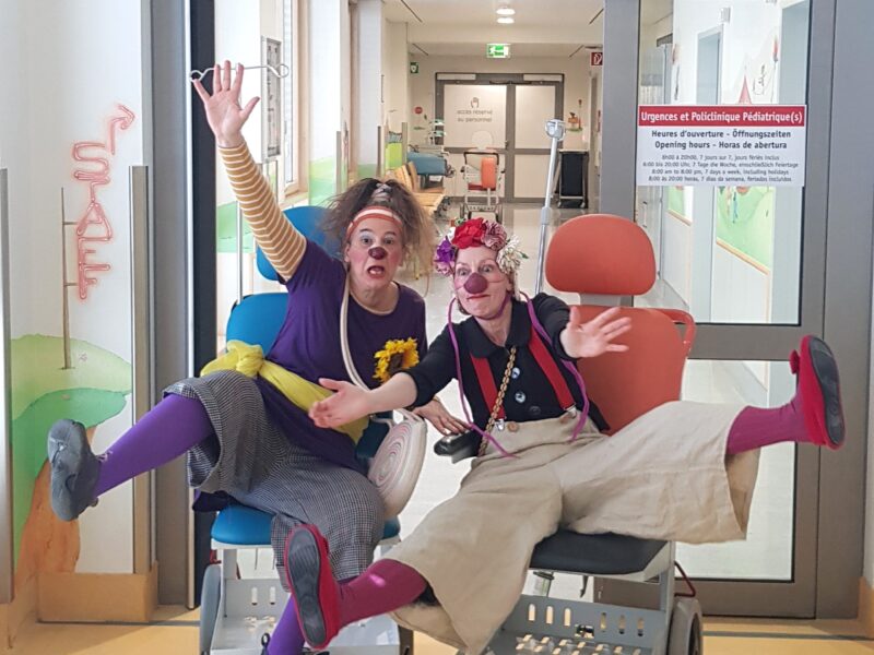 Deux clowns portant des tenues colorées et des nez rouges sont assis sur des chaises d'hôpital dans le couloir d'une clinique pédiatrique, faisant des signes et souriant avec des expressions exagérées. Au-dessus, un panneau indique en français qu'il s'agit d'une salle d'urgence pédiatrique.