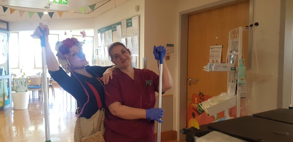Deux femmes souriant et posant avec des serpillières dans un couloir lumineux ; l'une porte une blouse médicale et des gants, l'autre des vêtements colorés et un bandeau de fleurs. Du matériel de nettoyage et des tableaux sont visibles à l'arrière-plan.