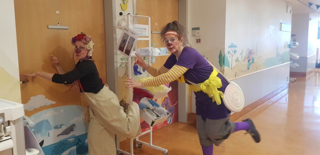 Deux adultes déguisés en clowns, avec des vêtements colorés et des visages peints, prennent des poses amusantes dans un couloir orné de peintures murales et d'équipements médicaux, créant ainsi une atmosphère amusante et animée.