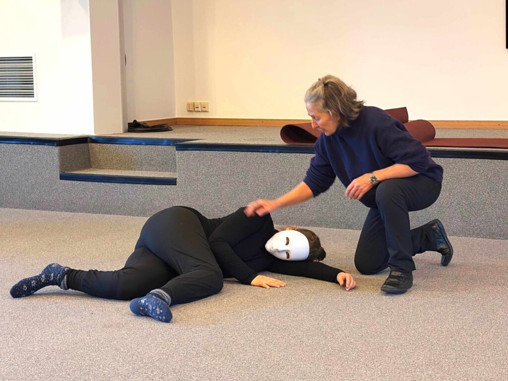 Une personne portant des vêtements noirs et un masque blanc est allongée sur le côté sur le sol, tandis qu'une autre personne s'agenouille à côté d'elle et lui touche doucement l'épaule dans un environnement intérieur bien éclairé.