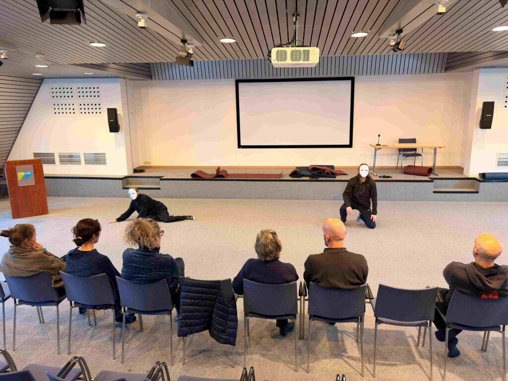 Deux personnes portant des masques blancs se produisent sur une scène devant un public assis de sept personnes. Les artistes sont au sol et prennent des poses dramatiques. Un grand écran blanc et un podium se trouvent à l'arrière-plan.