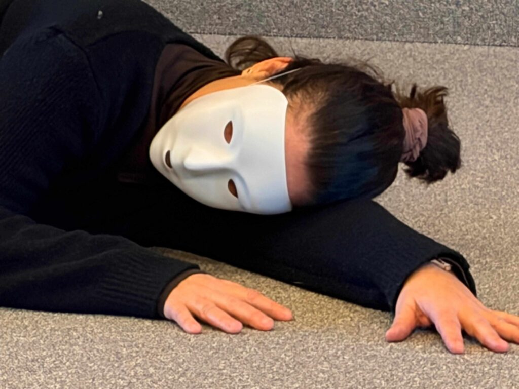 Une personne portant un masque blanc sans expression et des vêtements noirs est allongée sur un sol moquetté, la tête reposant sur son bras, face à la caméra.