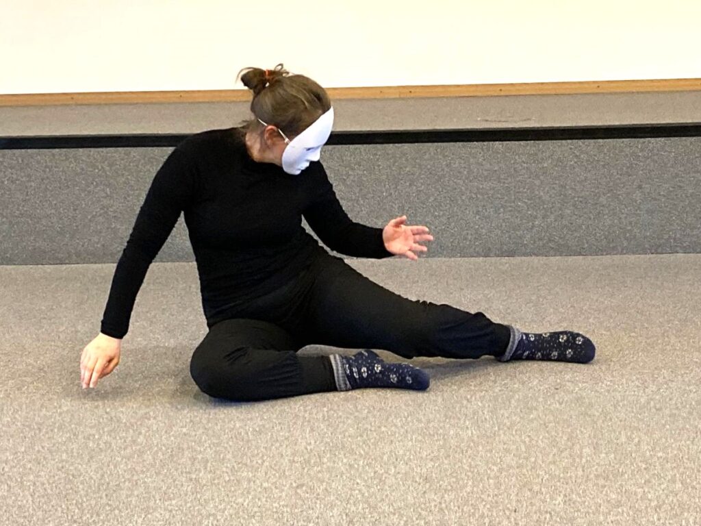 Une personne portant un masque blanc, une chemise noire et un pantalon noir est assise sur un sol moquetté, une jambe pliée et une jambe tendue, portant des chaussettes à motifs, et pose avec les bras tendus et la tête tournée sur le côté.