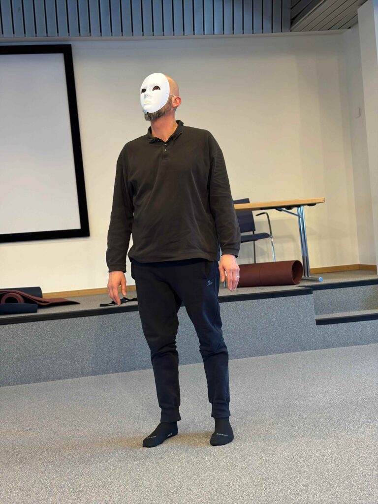 Une personne portant un masque blanc, une chemise noire à manches longues, un pantalon noir et des chaussettes se tient à l'intérieur, sur un sol moquetté, devant une scène équipée d'une table, de chaises et d'un grand écran blanc.