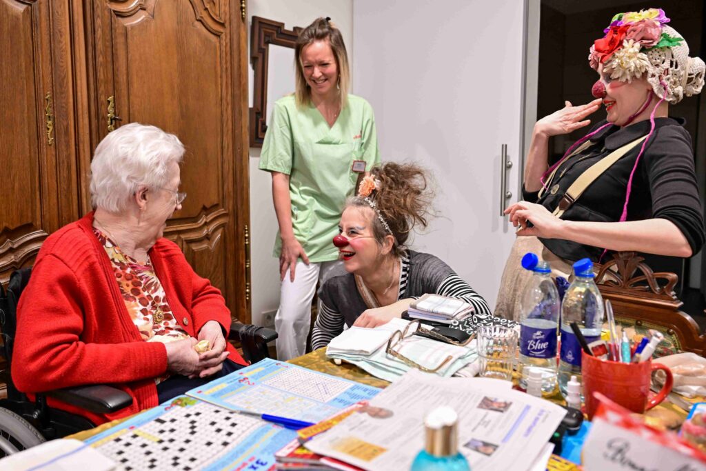 Une femme âgée en fauteuil roulant sourit à deux clowns au nez rouge qui la divertissent, sous le regard souriant d'une aide-soignante en blouse. La table est couverte de journaux, de bouteilles d'eau et de papiers.