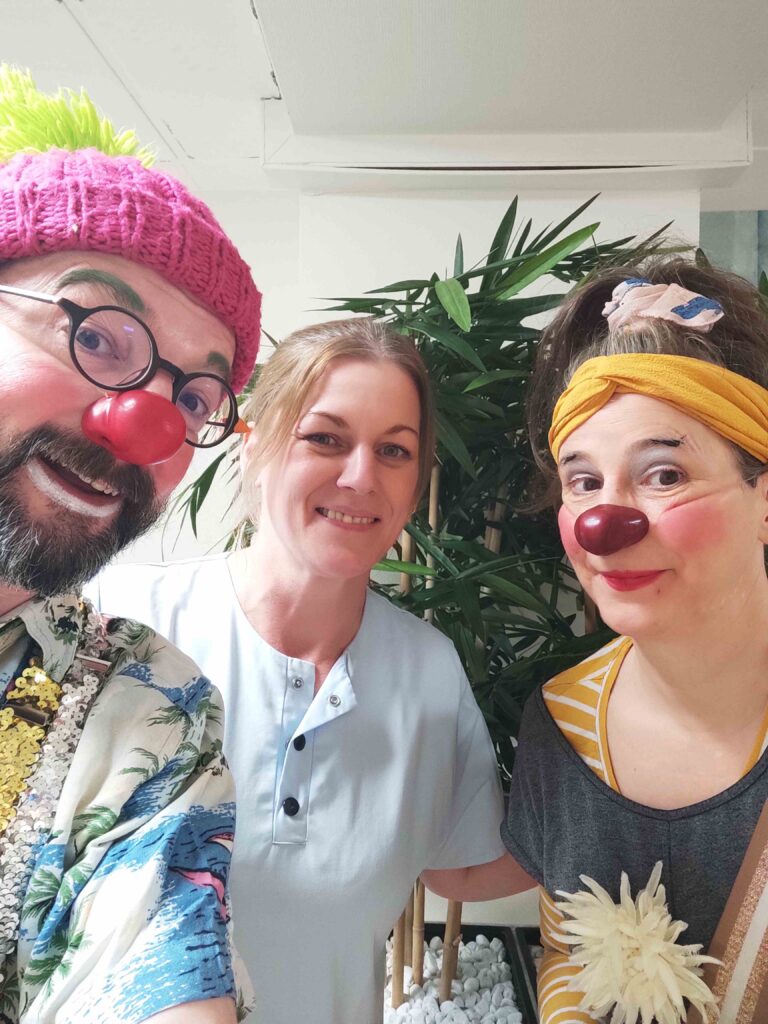 Trois adultes posent pour un selfie. Deux d'entre eux sont habillés en clowns avec un nez rouge et des accessoires colorés, tandis que la personne du milieu porte une chemise bleu clair et sourit. Une plante verte se trouve à l'arrière-plan.