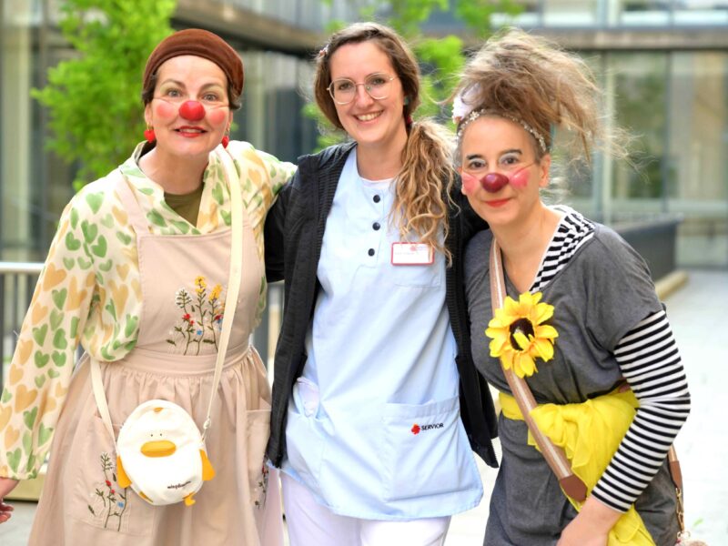 Trois femmes posent ensemble et sourient ; deux sont habillées comme des clowns avec des nez rouges, des vêtements colorés et des accessoires amusants, et l'autre porte un uniforme médical bleu clair avec un badge, en plein air dans un endroit lumineux.