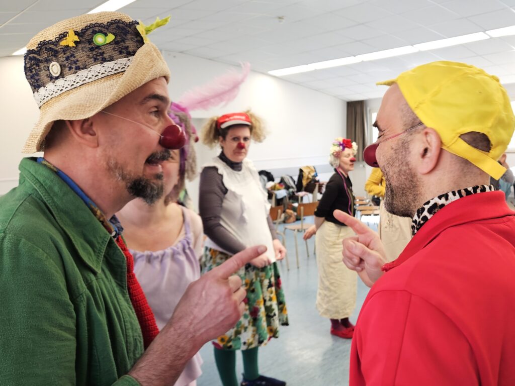 Deux personnes déguisées en clowns, avec des nez rouges et des costumes colorés, se sourient et se montrent du doigt dans une pièce très éclairée, tandis que d'autres personnes déguisées en clowns se tiennent à l'arrière-plan.