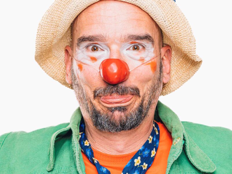 Un homme habillé en clown avec un nez rouge, de la peinture faciale blanche et orange, un chapeau de paille avec une plume jaune, une veste verte, une chemise orange et une cravate bleue à motifs fait une drôle de tête sur un fond blanc.