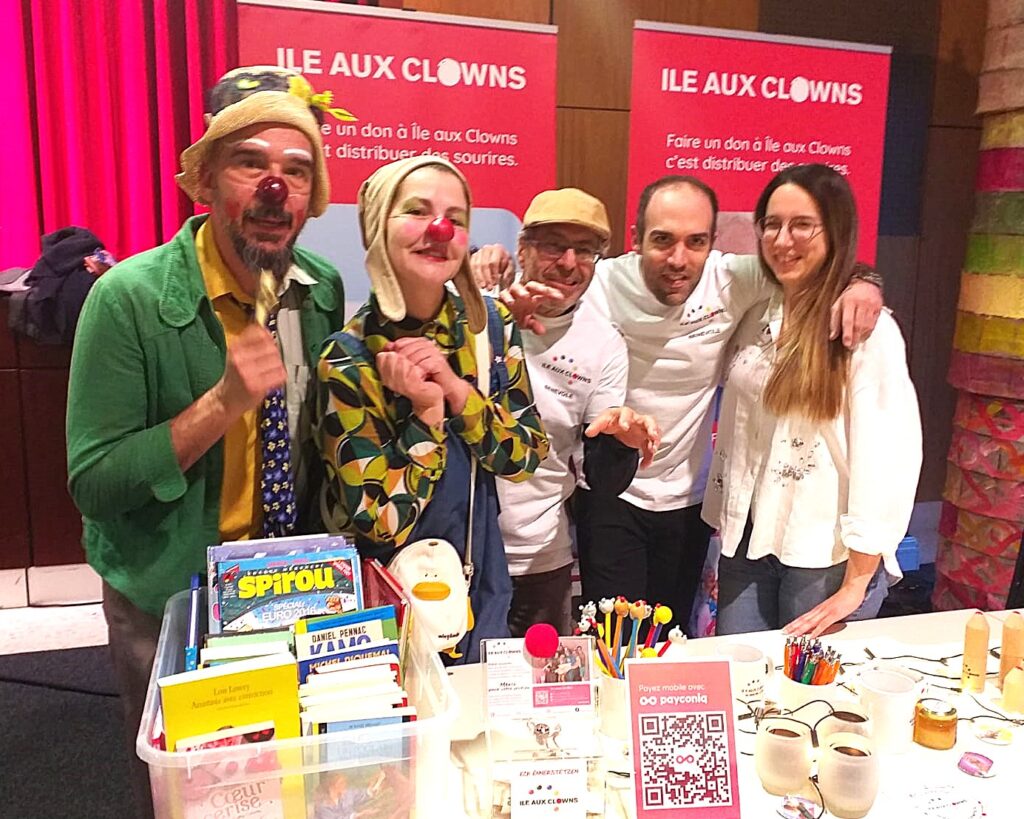 Cinq personnes, dont deux déguisées en clowns, sourient derrière une table où se trouvent des livres, des fournitures artistiques et une boîte à dons, lors d'une manifestation pour l'Ile aux Clowns, dont le nom figure sur des banderoles à l'arrière-plan.
