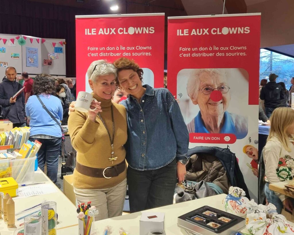 Deux femmes sourient et s'étreignent devant un stand de l'ILE AUX CLOWNS, une association caritative qui promeut le sourire, entourées de banderoles colorées, de boîtes de dons et d'objets artisanaux. Des visiteurs et des enfants flânent dans cette manifestation très animée qui se déroule en intérieur.