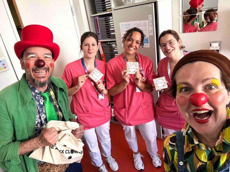 Deux personnes déguisées en clowns posent aux côtés de trois membres du personnel soignant souriants, vêtus d'uniformes roses et tenant de petits paquets cadeaux. Ils se trouvent tous ensemble dans une chambre d'hôpital lumineuse.