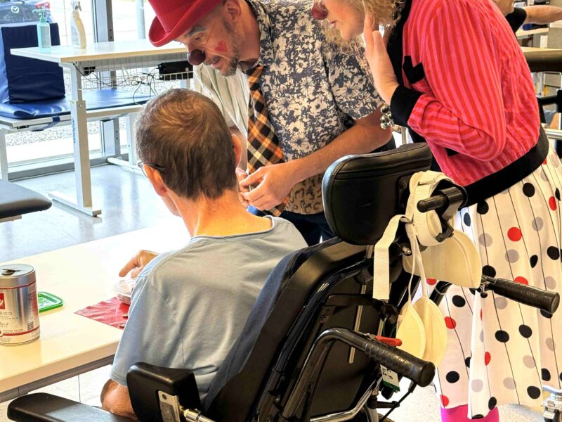 Deux clowns vêtus de costumes colorés discutent avec une personne en fauteuil roulant, assise à une table dans une pièce lumineuse, créant ainsi une ambiance joyeuse et chaleureuse. On aperçoit d'autres personnes et de grandes fenêtres en arrière-plan.