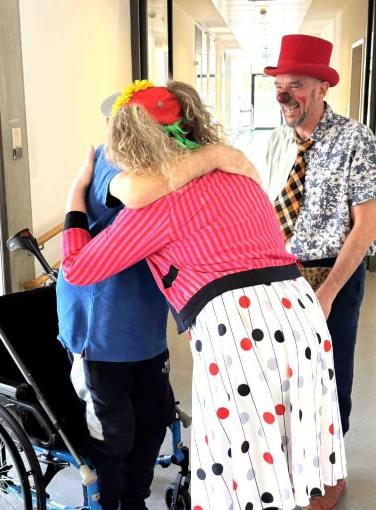 Une femme déguisée en clown, avec un chapeau rouge et une jupe à pois, serre dans ses bras un enfant en fauteuil roulant. Un autre homme, lui aussi déguisé en clown avec un chapeau rouge et le visage peint, sourit à proximité dans un couloir lumineux.