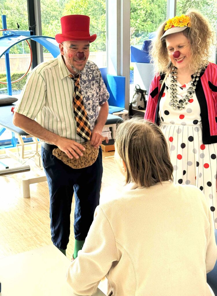 Deux personnes déguisées en clowns, l'une coiffée d'un chapeau rouge et vêtue d'habits dépareillés, l'autre portant une couronne de fleurs et une robe à pois, sourient et discutent avec une personne assise dans une pièce lumineuse.