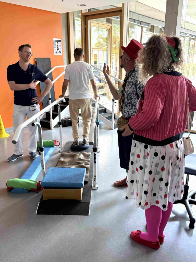 Deux personnes déguisées en clowns discutent avec un professionnel de santé tandis qu'une autre marche sur des barres parallèles dans une salle de rééducation. On aperçoit des tapis de rééducation et du matériel de rééducation au sol.