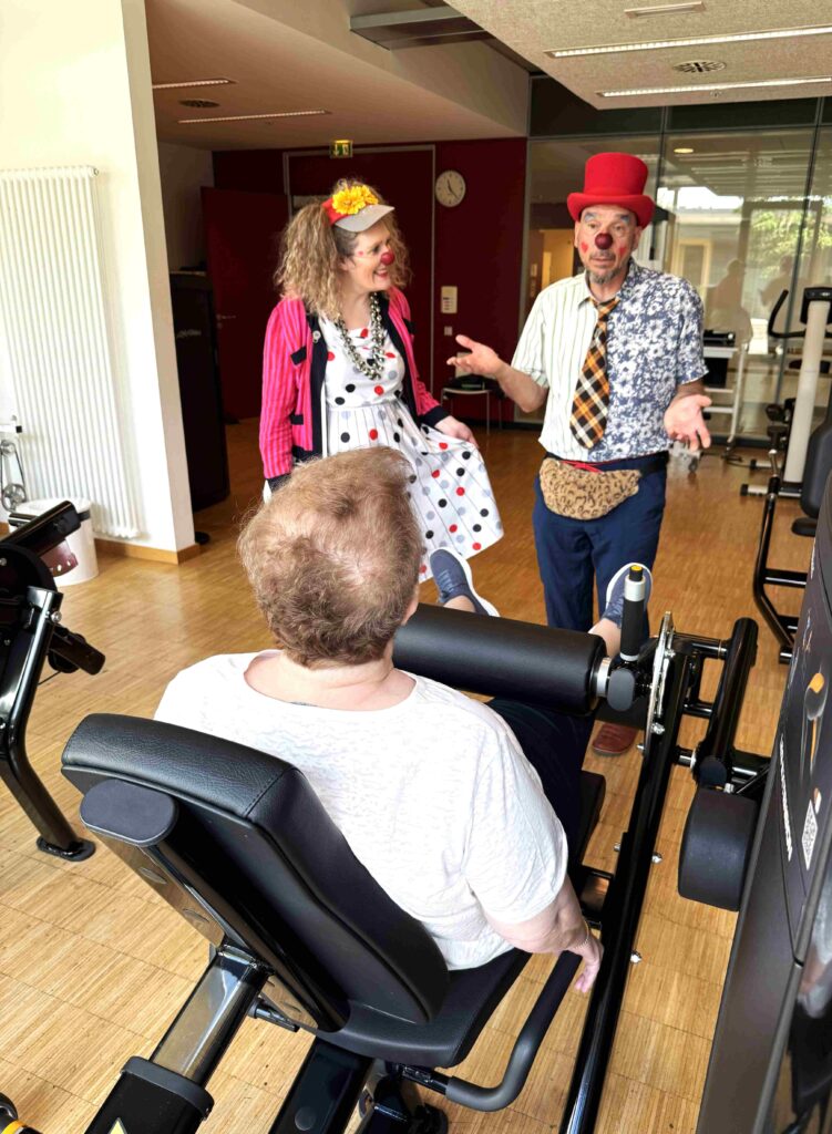Deux clowns, vêtus de costumes colorés et maquillés, discutent joyeusement avec une personne qui s'entraîne sur une machine à cuisses dans une salle de sport, créant ainsi une ambiance ludique et détendue.