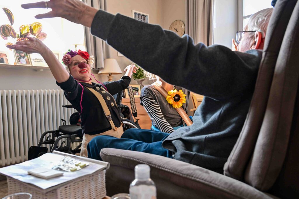 Une femme déguisée en clown, avec un nez rouge, des bretelles et une couronne de fleurs, divertit des personnes âgées assises dans un salon chaleureux, les faisant rire et sourire grâce à ses gestes amples et à son énergie enjouée.
