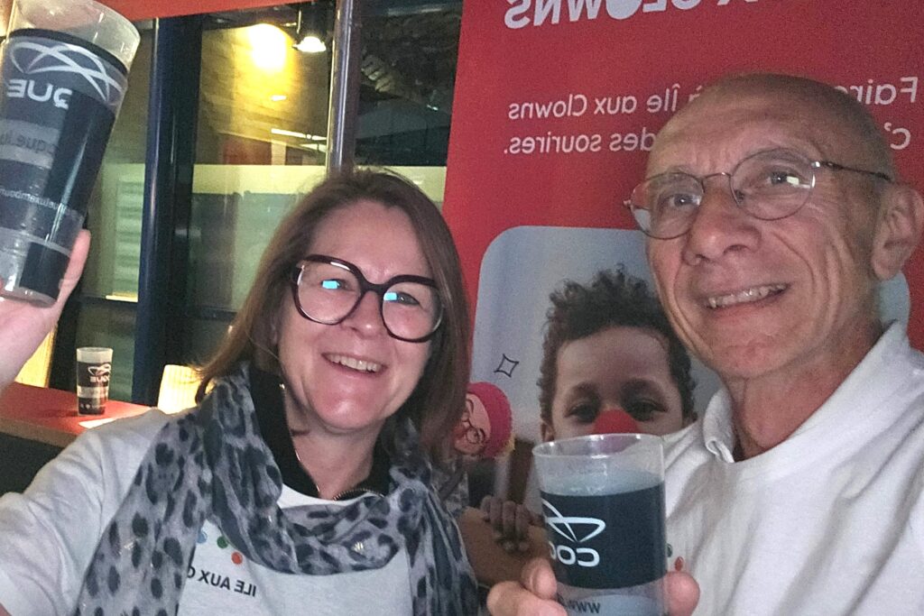 Deux personnes souriantes portant des lunettes brandissent des gobelets en plastique lors d'un événement organisé en salle. Derrière elles, on aperçoit une grande banderole rouge avec un texte en blanc et le visage d'un enfant partiellement visible. L'ambiance semble joyeuse et conviviale.