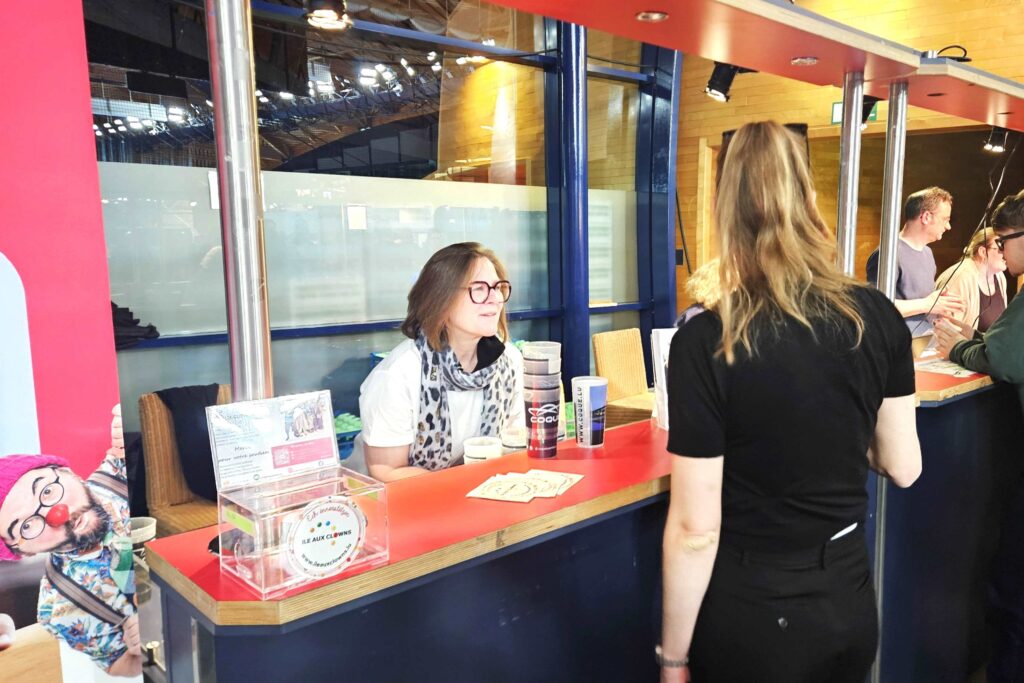 Une femme à lunettes est assise à un comptoir ; elle sourit et discute avec une autre femme qui se tient debout devant elle. Des tracts, des badges et un présentoir sont posés sur le comptoir. À l'arrière-plan, plusieurs personnes discutent aux comptoirs voisins.