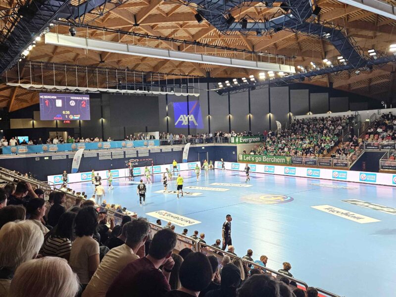 Une salle comble où les spectateurs assistent à un match de handball ; les joueurs en pleine action sur un terrain bleu, un tableau d'affichage électronique indiquant 1-2, ainsi que des banderoles et des publicités tout autour de la salle.