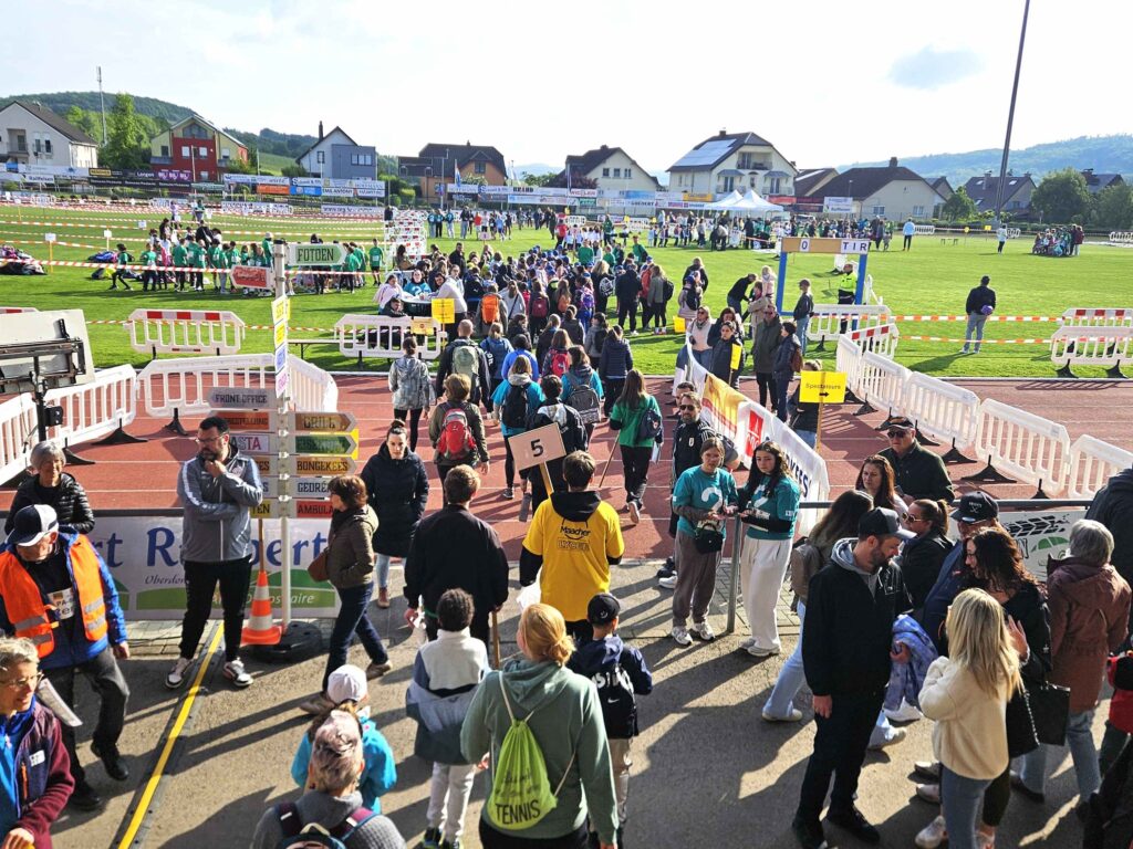 Une foule se rassemble à l'occasion d'un événement sportif en plein air sur un terrain. Les gens franchissent les barrières, portent des maillots numérotés et des cordons d'identification pour l'événement. Des tentes, des enseignes et des maisons sont visibles au loin sous un ciel partiellement nuageux.