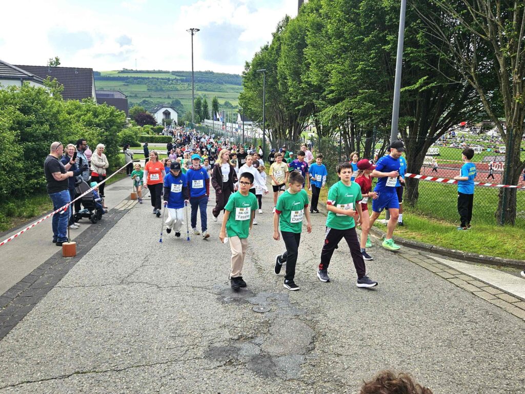 Un groupe d'enfants, portant des dossards numérotés et des chemises colorées, marchent et courent le long d'une rue lors d'une course, entourés de spectateurs et d'arbres, avec un paysage vallonné en arrière-plan.
