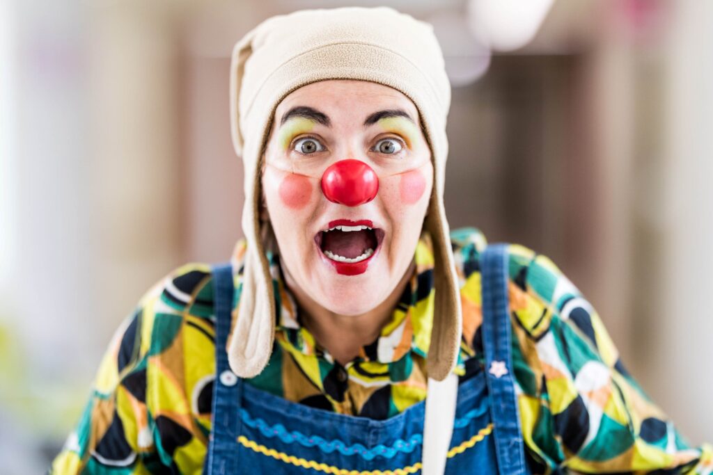 Une personne habillée en clown, avec un nez rouge, un visage peint en couleurs, un chapeau ludique et une chemise à motifs, sourit largement avec une expression enthousiaste.