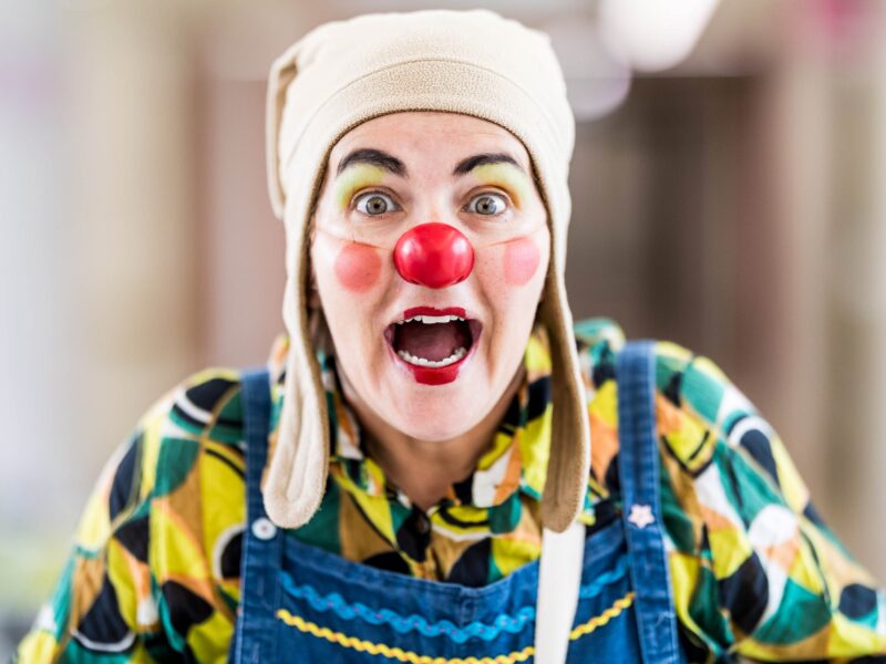 Une personne habillée en clown, avec un nez rouge, un visage peint en couleurs, un chapeau ludique et une chemise à motifs, sourit largement avec une expression enthousiaste.
