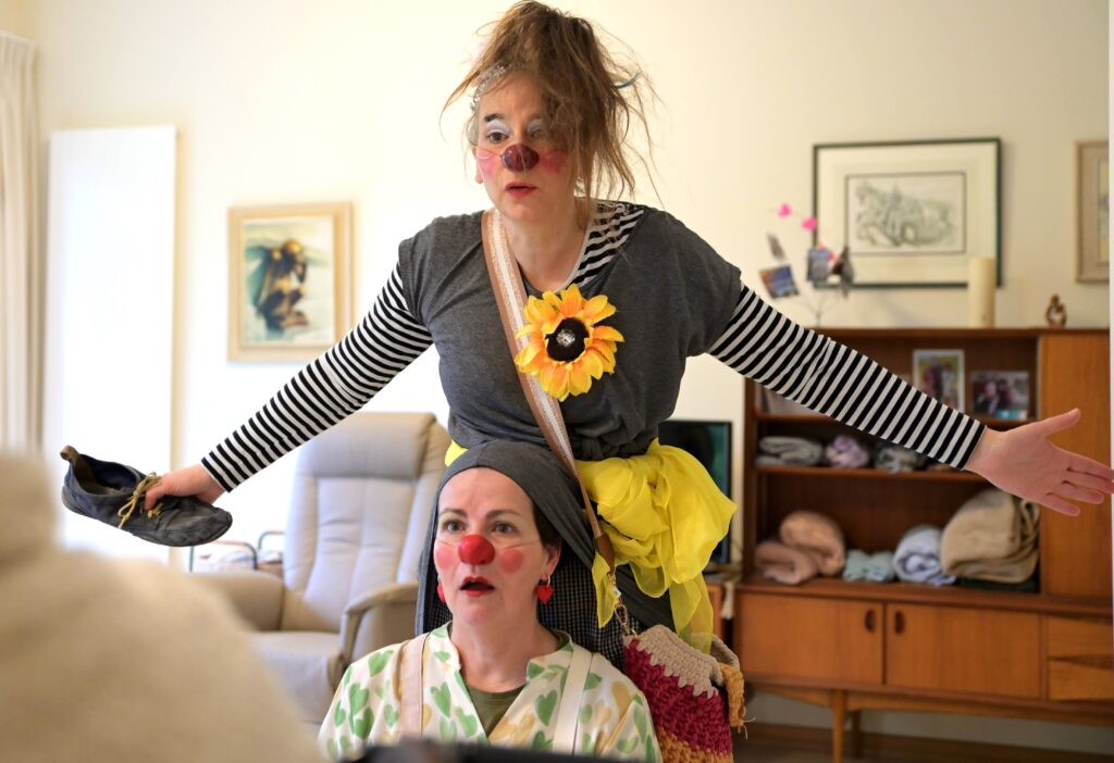 Deux femmes déguisées en clowns se produisent à l'intérieur. L'une se tient derrière l'autre, les bras tendus, tenant une chaussure, tandis que la femme assise a l'air surprise. Les deux femmes portent des nez rouges et des costumes colorés.