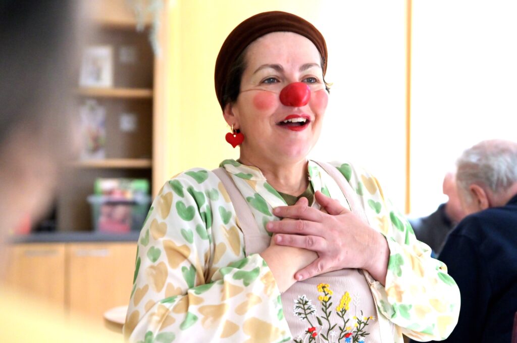 Une femme portant un nez de clown rouge, des boucles d'oreilles en forme de cœur et une chemise à motif de cœur sourit chaleureusement en portant ses mains à sa poitrine dans une pièce très éclairée avec d'autres personnes en arrière-plan.