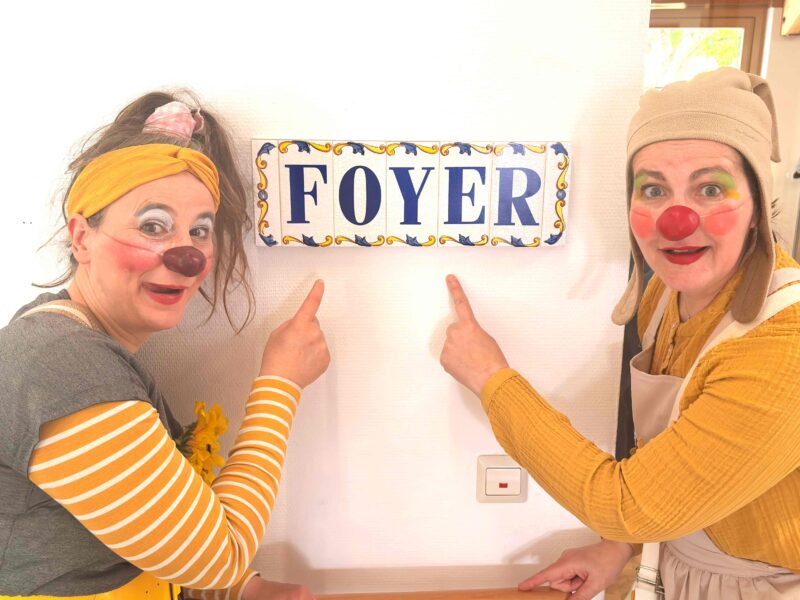 Deux femmes déguisées en clowns, portant des nez rouges et des costumes colorés, sourient et pointent du doigt un panneau décoratif indiquant FOYER sur un mur blanc. Une lumière vive traverse une fenêtre à l'arrière-plan.