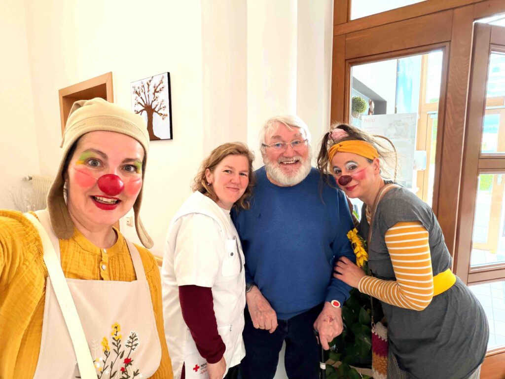 Quatre adultes se tiennent ensemble en souriant. Deux femmes sont habillées comme des clowns avec des nez rouges et de la peinture faciale, tandis que l'autre femme porte un uniforme médical blanc. Un homme plus âgé avec une barbe se tient au centre. Ils sont à l'intérieur, près d'une porte.