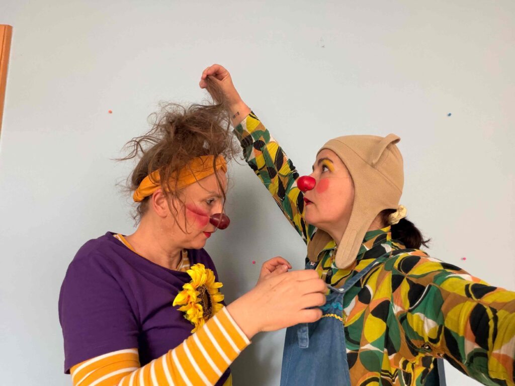 Deux personnes déguisées en clowns se font face ; l'une porte un bandeau jaune et une chemise violette et a les cheveux en désordre, tandis que l'autre, vêtue d'une chemise à motifs et d'un chapeau beige, lui soulève les cheveux. Tous deux ont le nez rouge et des expressions animées.