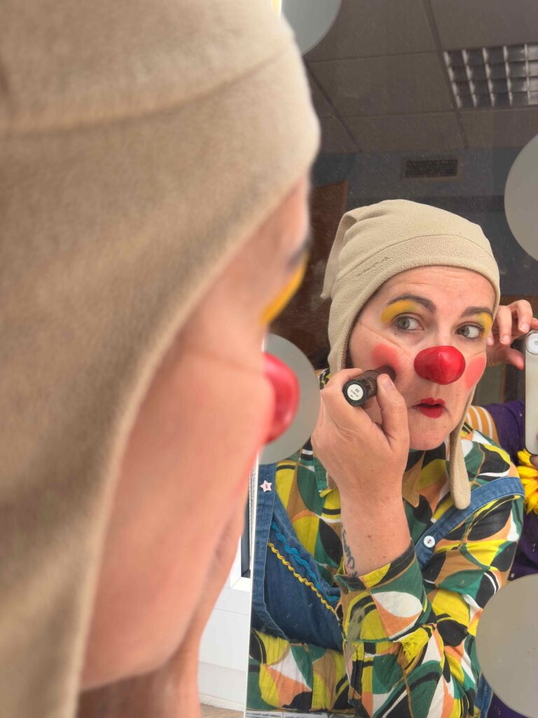 Une personne vêtue de vêtements colorés et d'un chapeau beige se maquille le visage et se met du fard à joues rouge en se regardant dans un miroir. Elle a déjà un nez de clown rouge et des yeux jaunes.