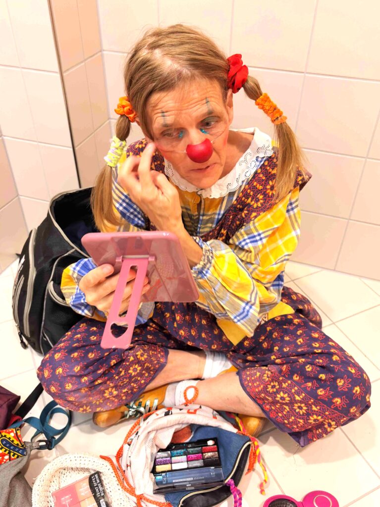 Une personne déguisée en clown est assise, les jambes croisées, et se maquille à l'aide d'un petit miroir. Elle porte des nattes, un nez rouge et des vêtements colorés. Des articles de maquillage et des accessoires sont éparpillés autour d'elle sur le sol.