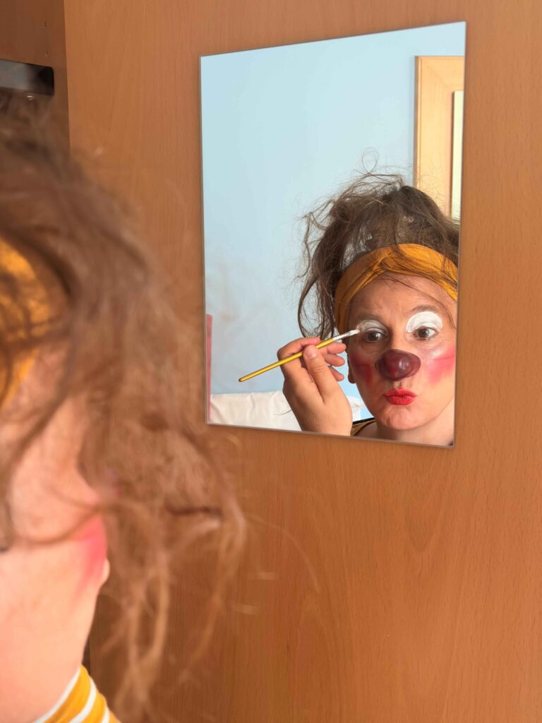Une personne se regarde dans un miroir tout en se maquillant le visage au pinceau avec un maquillage de clown, comprenant des cernes blancs et un nez rouge. Son reflet montre une expression enjouée et un bandeau jaune dans des cheveux en désordre.