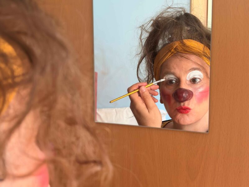Une personne se regarde dans un miroir tout en se maquillant le visage au pinceau avec un maquillage de clown, comprenant des cernes blancs et un nez rouge. Son reflet montre une expression enjouée et un bandeau jaune dans des cheveux en désordre.