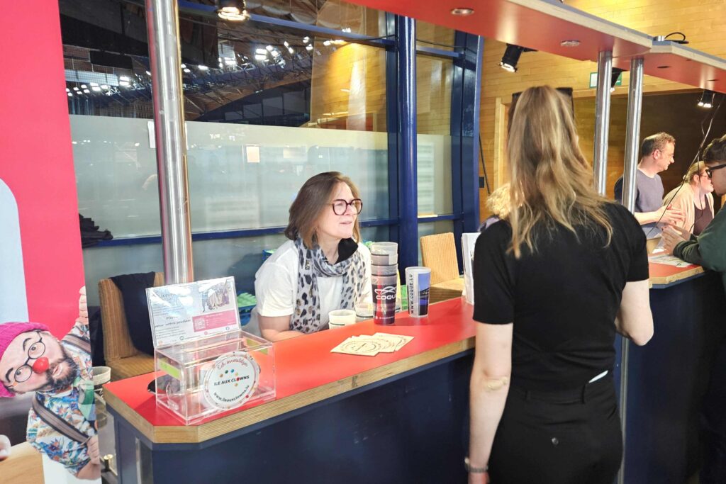 Une femme à lunettes est assise derrière un comptoir bleu et parle à une femme debout. Des supports promotionnels et une silhouette découpée représentant un homme barbu se trouvent sur le comptoir. D'autres personnes sont visibles à l'arrière-plan.