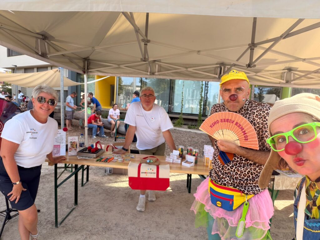 Quatre adultes posent devant un stand de marché en plein air. Deux d'entre eux portent des chemises blanches, tandis que les deux autres sont vêtus de costumes colorés et fantaisistes, avec des perruques, des chapeaux et du maquillage. La table, placée sous un auvent, est garnie d'objets artisanaux et de brochures.