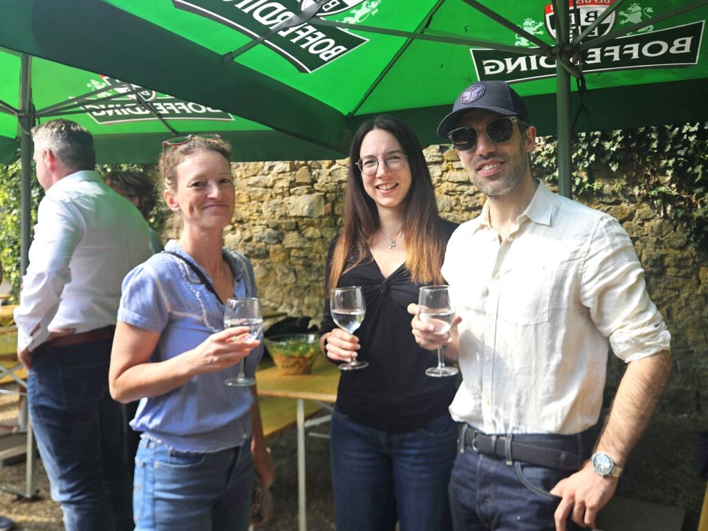 Trois personnes se tiennent à l'extérieur sous des parasols verts, souriantes, un verre de vin à la main. On aperçoit un mur en pierre et une autre personne à l'arrière-plan. Cela ressemble à une réunion entre amis dans une ambiance décontractée.