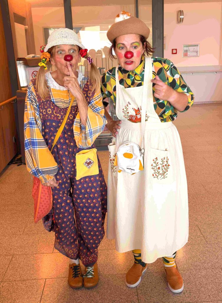 Deux adultes déguisés en clowns, avec un nez rouge, des vêtements colorés, des chapeaux et des accessoires amusants, se tiennent à l'intérieur. L'un d'eux porte un doigt à ses lèvres, tandis que l'autre pointe du doigt vers l'avant. Tous deux ont des visages espiègles et expressifs.
