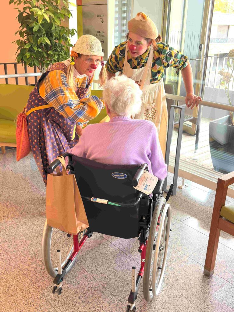 Deux clowns discutent avec une femme âgée en fauteuil roulant dans une pièce lumineuse. L'un des clowns se penche vers elle, tandis que l'autre se tient à côté ; tous deux sourient et portent des costumes colorés ainsi qu'un nez rouge.