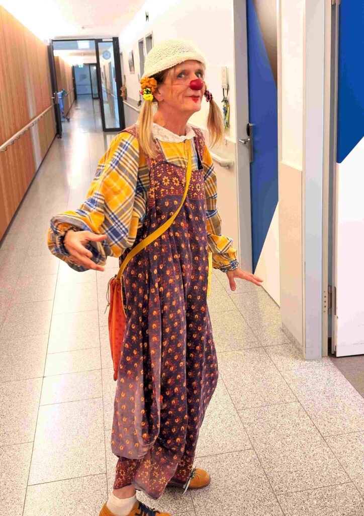 Une personne déguisée en clown, avec des nattes, un chapeau blanc, une chemise à carreaux, un pull à motifs et un nez rouge, se tient dans un couloir, tenant quelque chose dans une main et souriant.