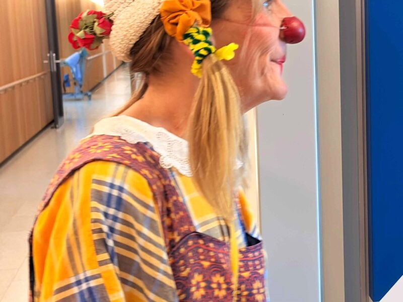 Une personne déguisée en clown, coiffée d'un bonnet en tricot blanc, d'un nez de clown rouge, de chouchous colorés et vêtue d'un costume à motifs, se tient dans un couloir, souriante, le regard tourné vers la droite.