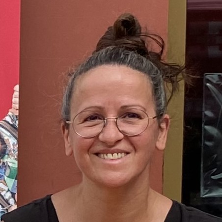 Une femme aux cheveux châtains, portant des lunettes et coiffée d'un chignon haut, sourit à la caméra. Elle porte un haut noir et se tient debout devant un fond rouge et marron.